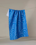 The Sareka Unique "High Tide" tea towel, featuring a blue and white abstract swirl design and made from ultra-absorbent recycled materials, hangs on a clothesline against a pale background.