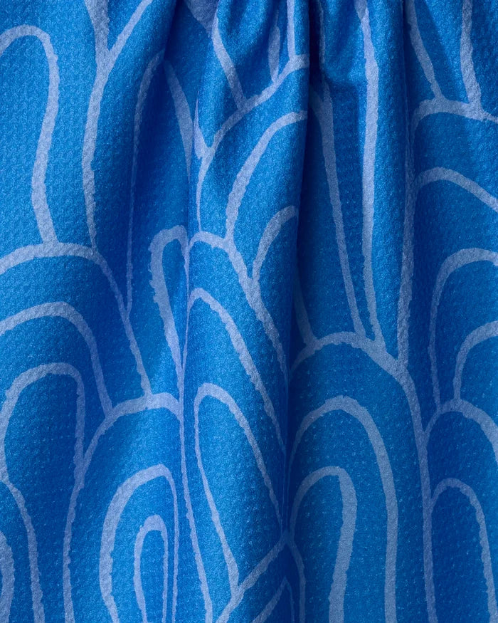 A close-up of the Sareka Unique "High Tide" tea towel shows blue recycled fabric with a light blue wavy pattern, gathered ruching at the top, and a slightly pebbled texture.