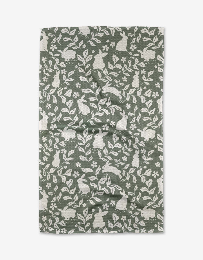 The Hippity Hop tea towel by Charlie Rowan Designs features a repeating pattern of white rabbits, leaves, and flowers in green and white on slightly wrinkled recycled fabric, laid flat on a white background.