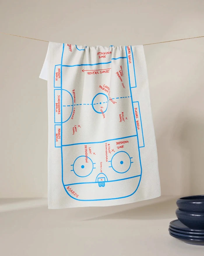 The GEOMETRY Hockey kitchen towel, in white, hangs on a line and displays a blue and red ice hockey rink diagram with labeled zones and markings; dark blue bowls sit in the lower right corner.