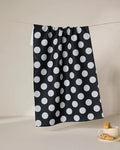The GEOMETRY Hole In One ultra-absorbent tea towel, featuring large white polka dots on black, hangs on a clothesline; nearby, a white cup with saucer and cookies highlights the towel’s perfect mix of charm and practicality.
