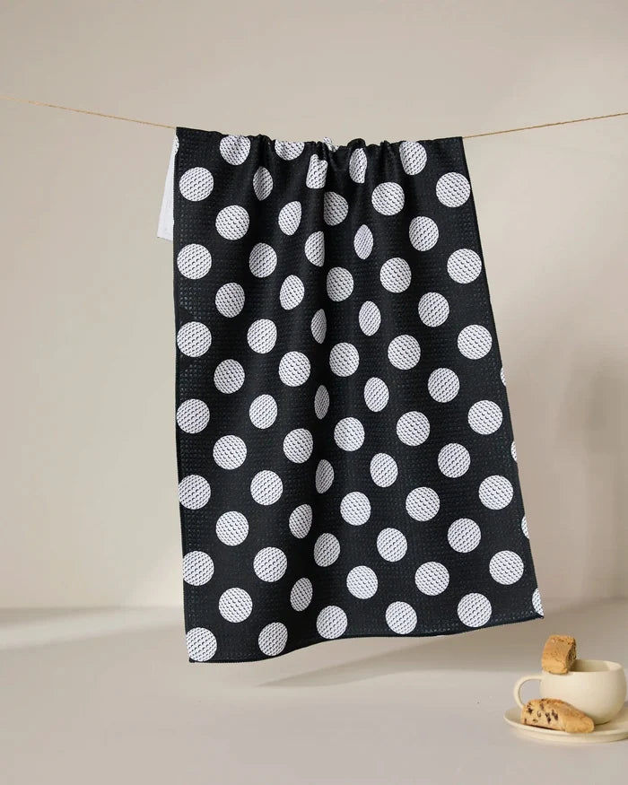 The GEOMETRY Hole In One ultra-absorbent tea towel, featuring large white polka dots on black, hangs on a clothesline; nearby, a white cup with saucer and cookies highlights the towel’s perfect mix of charm and practicality.