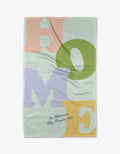 The Home tea towel by Quotes by Christie features large pastel letters spelling HOME in pink, green, purple, and yellow on recycled fabric, with “Is Wherever My People Are” in small blue cursive underneath.