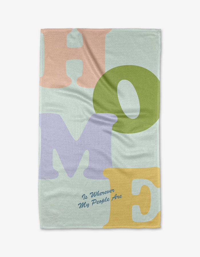 The Home tea towel by Quotes by Christie features large pastel letters spelling HOME in pink, green, purple, and yellow on recycled fabric, with “Is Wherever My People Are” in small blue cursive underneath.