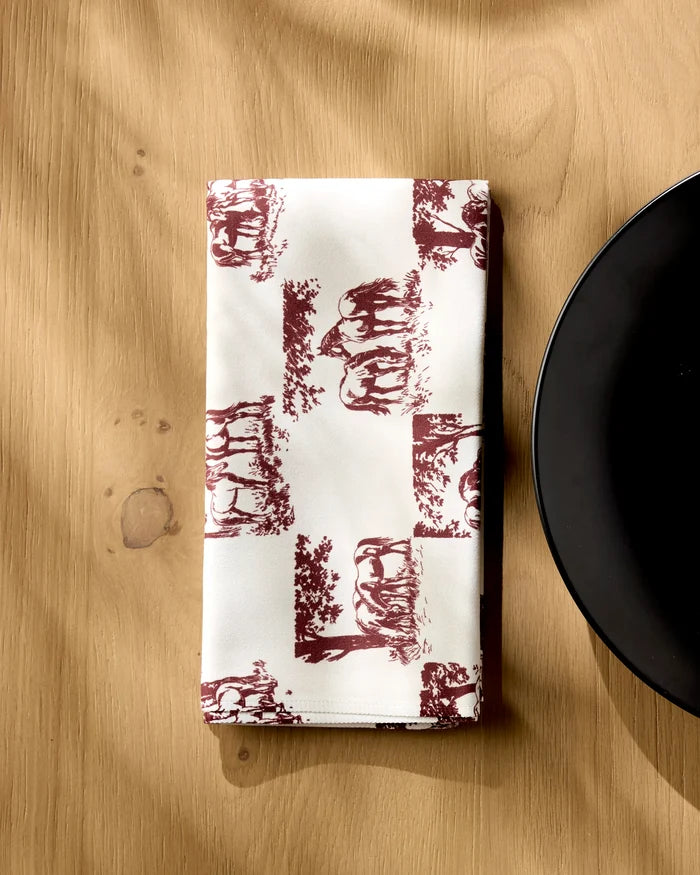 A folded Geometry Horse Check Cream napkin with burgundy animal and tree illustrations sits on a light wooden table near the right edge of a round black plate.