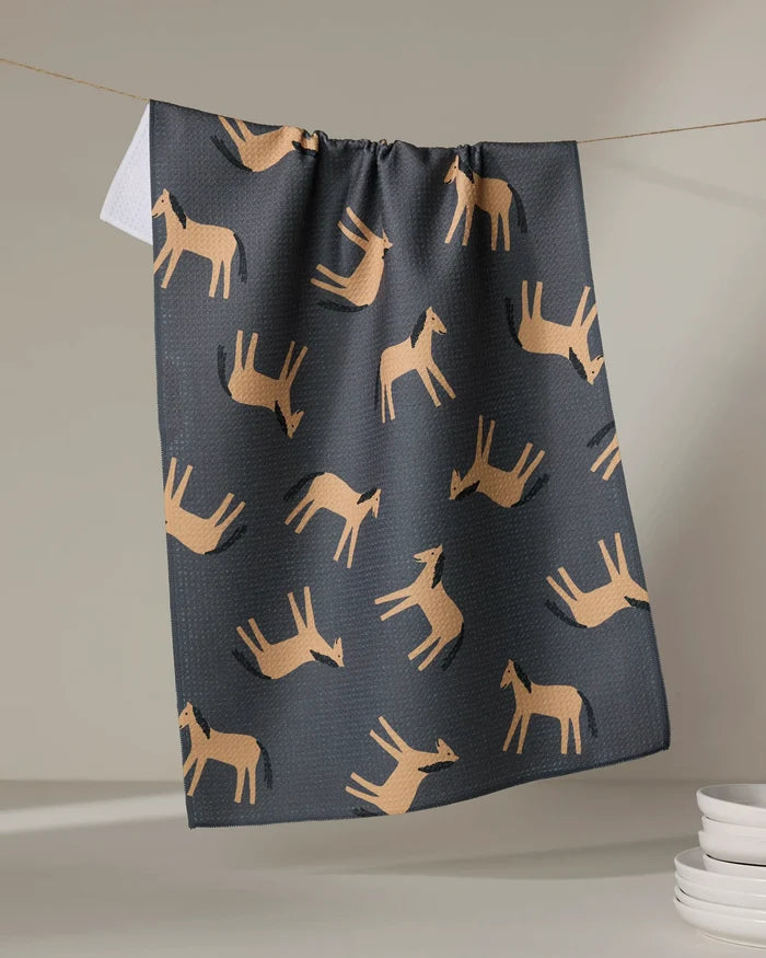 The "Horses" tea towel by Coit Creative, featuring a repeating tan horse pattern, hangs above white plates against a neutral background.