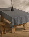 A rectangular wooden table is draped with Geometry’s Houndstooth Black tablecloth, topped with a small round black vase holding dry branches. A wooden stool sits beside the table on a light wooden floor.
