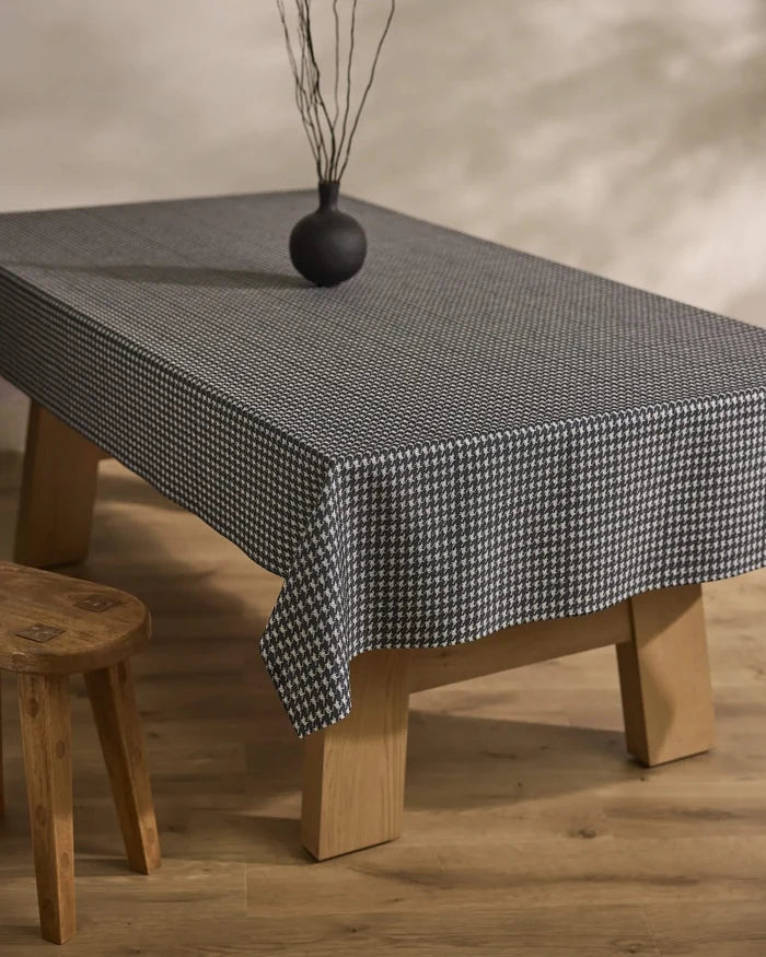 A rectangular wooden table is draped with Geometry’s Houndstooth Black tablecloth, topped with a small round black vase holding dry branches. A wooden stool sits beside the table on a light wooden floor.