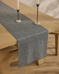 A wooden dining table is topped with the Geometry Houndstooth Black stain-resistant table runner, accented by two brass candlesticks with white candles, all placed on a light wood floor.