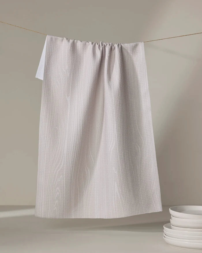 The Houston Wood tea towel by Kailee Wright, featuring a light gray textured pattern and made from recycled materials, hangs on a string with a stack of white plates in the lower right corner against a neutral background.