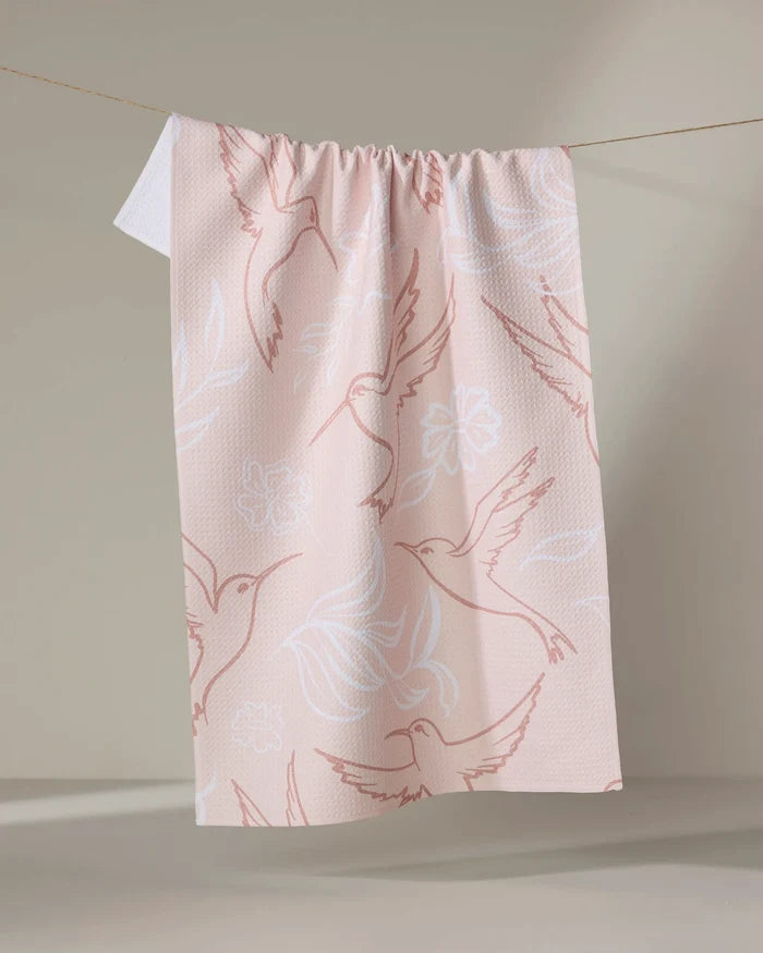 The 'Hummingbirds' tea towel by Elena Fay features a pink design with white and dark pink hummingbird and flower outlines, hanging from a line against a neutral background.