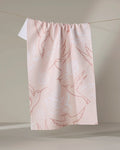 The 'Hummingbirds' tea towel by Elena Fay features a pink design with white and dark pink hummingbird and flower outlines, hanging from a line against a neutral background.