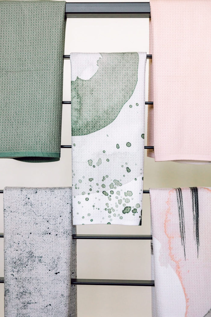 Five Hunter Abstract towels by Kailee Wright hang on a black rack against a beige wall. Made from recycled materials, the set includes green textured, pink textured, white with green splatters, gray speckled, and white with abstract brushstrokes.