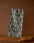 The Geometry Husk bar towel, featuring a black and gray leaf pattern and made from recycled materials, hangs on a clothesline with clothespins. In front, a loaf of bread sits on a wooden board against a brown backdrop.