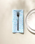 A black fork rests on a folded blue and white Hydrangea-patterned absorbent napkin by GEOMETRY, beside a white textured plate on a light beige tablecloth.