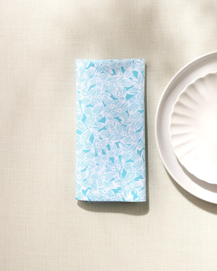 A GEOMETRY Hydrangea blue absorbent napkin with a white leaf pattern is placed beside a white scalloped plate on a textured beige surface.