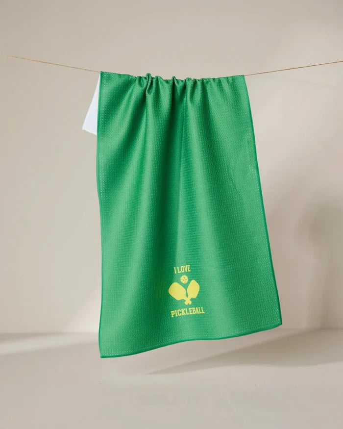 The GEOMETRY I Love Pickleball green tea towel, made from recycled materials, hangs on a line with a yellow graphic of two crossed pickleball paddles, a ball, and “I LOVE PICKLEBALL” near the bottom center.