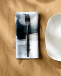 A black fork rests on Jennifer Janesko’s Icelandic Elements eco-friendly, abstract-patterned napkin beside a white square plate on a light wooden table, soft natural light casting gentle shadows.
