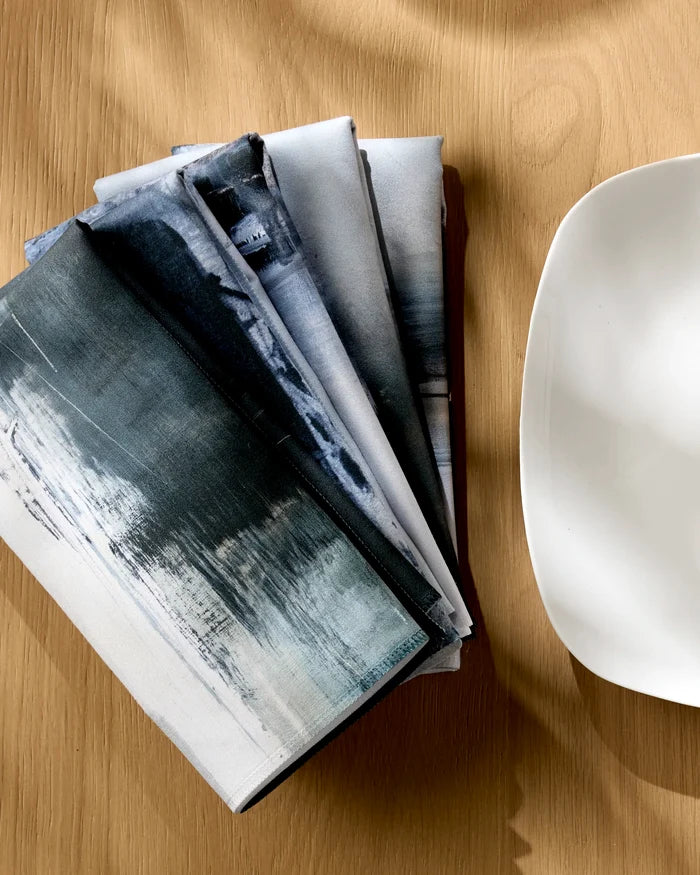 Four "Icelandic Elements" reusable dinner napkins by Jennifer Janesko, featuring abstract blue and gray patterns, are arranged fan-like on wood beside a white plate. Sunlight highlights this elegant, eco-friendly scene.
