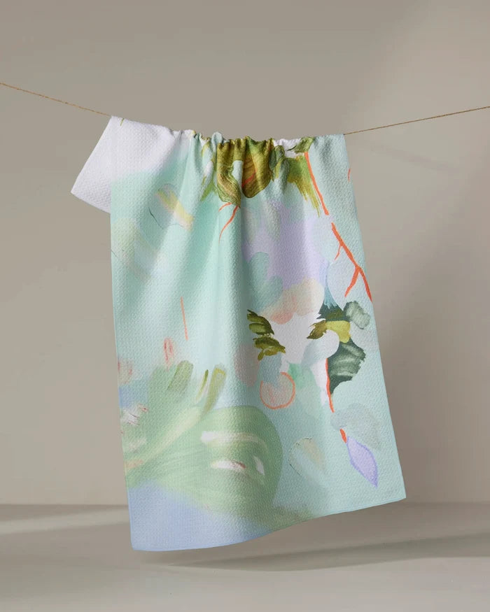 The "If I Were An Orchard" tea towel by Kristina Baker, featuring a pastel abstract leafy and floral pattern, hangs absorbently on a clothesline against a light background.