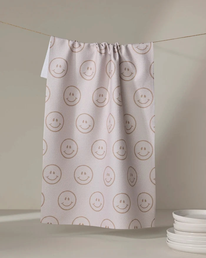 The "I'm Blushing" tea towel by Kelsi Fullmer, featuring cheerful smiley faces, hangs on a line to dry. A small stack of white plates sits in the lower right corner against a neutral background.