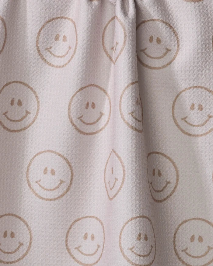 The "I'm Blushing" tea towel by Kelsi Fullmer is made from recycled materials and features a light-colored, textured fabric with brown smiley faces in circles. Soft folds form where the top is slightly gathered for added charm.