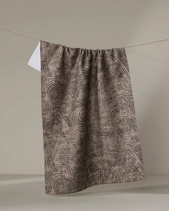 The "Imprint" kitchen towel by Studio Jeannot, featuring a brown and black abstract spiral pattern, hangs on a clothesline against a neutral background with a small white tag at the top left corner.