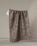 The "Imprint" kitchen towel by Studio Jeannot, featuring a brown and black abstract spiral pattern, hangs on a clothesline against a neutral background with a small white tag at the top left corner.