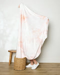 A person, wrapped in the large, light "In the Clouds" blanket by Geometry (except for their lower legs and white sneakers), stands behind a woven basket beside a wooden stool on a wooden floor.