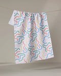 The "In the Moment" tea towel by Happy Hemera features a white textured fabric with colorful squiggly lines in blue, red, yellow, and green, hanging on a clothesline against a neutral backdrop.