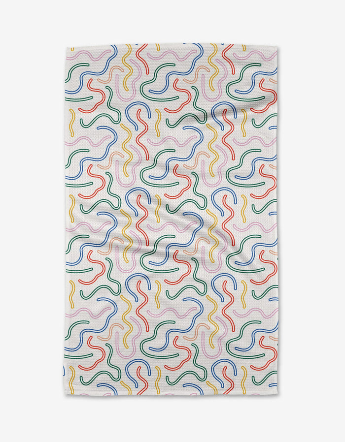 The In the Moment kitchen towel by Happy Hemera features a white fabric with wavy, multicolored squiggle patterns in red, yellow, green, and blue. Subtle creases are visible across the surface.