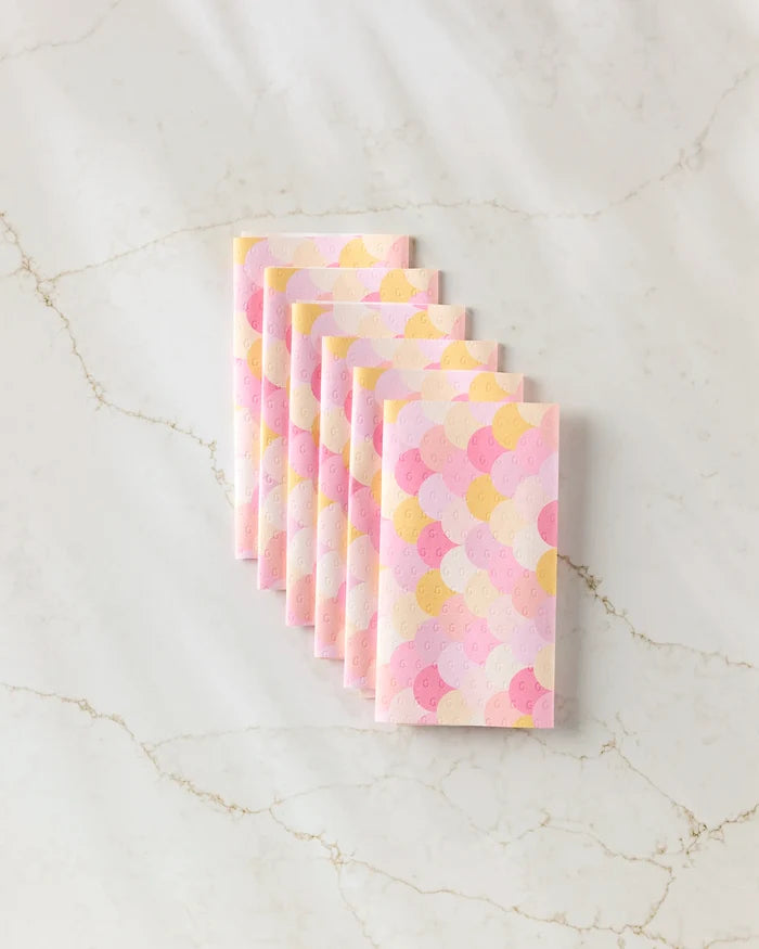 Eight eco-friendly GEOMETRY Inherit kitchen towels in pastel pink, yellow, and light purple scallop patterns are stacked neatly on a white marble surface with delicate gray and gold veining.