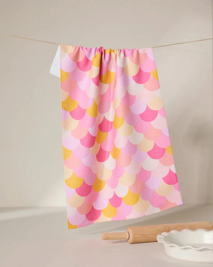 The GEOMETRY Inherit dish towel, featuring a pastel scallop pattern in pink, yellow, and orange, hangs on a string. A rolling pin and white ceramic dish sit on the white surface in the foreground.