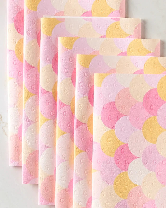Six folded reusable paper towels from GEOMETRY's Inherit collection, featuring a pastel scallop pattern in pink, yellow, and peach tones, are arranged in a fan on a white surface. Each towel has a subtle embossed letter G.