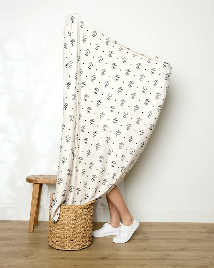 A person holds up the large, cream-colored Inked Bloom blanket by Cole Strem, featuring a black floral pattern. Only their lower legs and white sneakers show as they stand beside a wooden stool and woven basket on a wooden floor.
