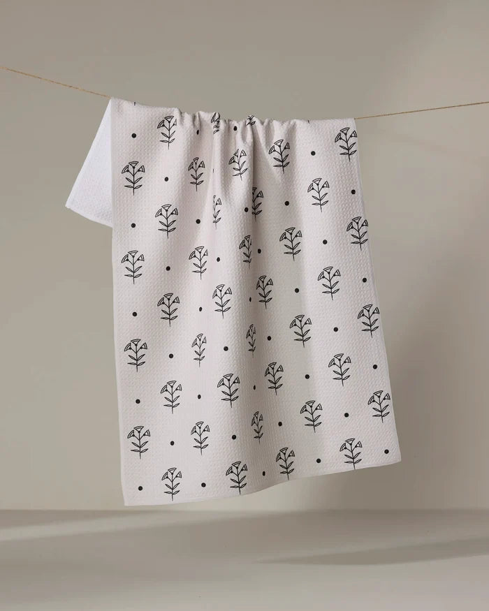 The Cole Strem "Inked Bloom" dish towel, featuring a black floral and dot pattern on white, is shown hanging over a clothesline against a neutral background.