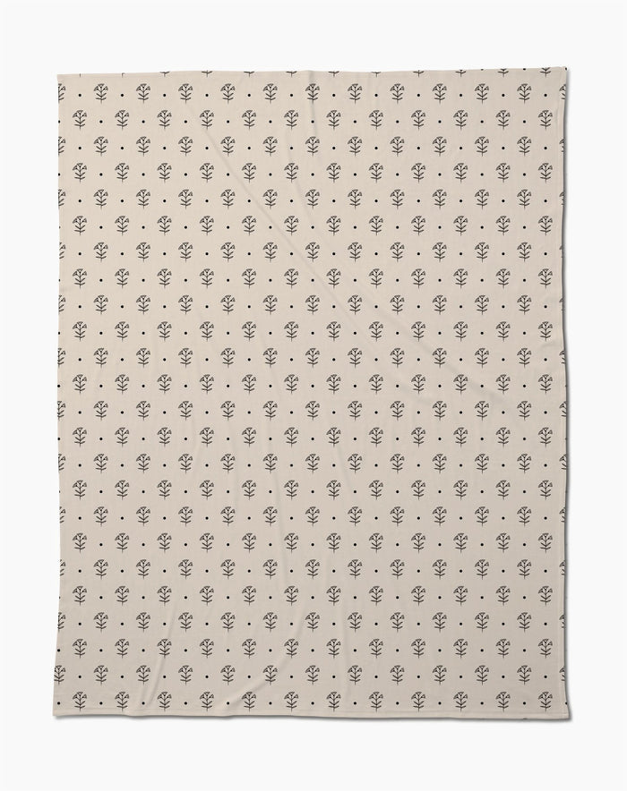 The Inked Bloom by Cole Strem is an eco-friendly, beige blanket with a simple, repeating black floral motif. Made from recycled materials, it appears soft with subtle natural wrinkles.
