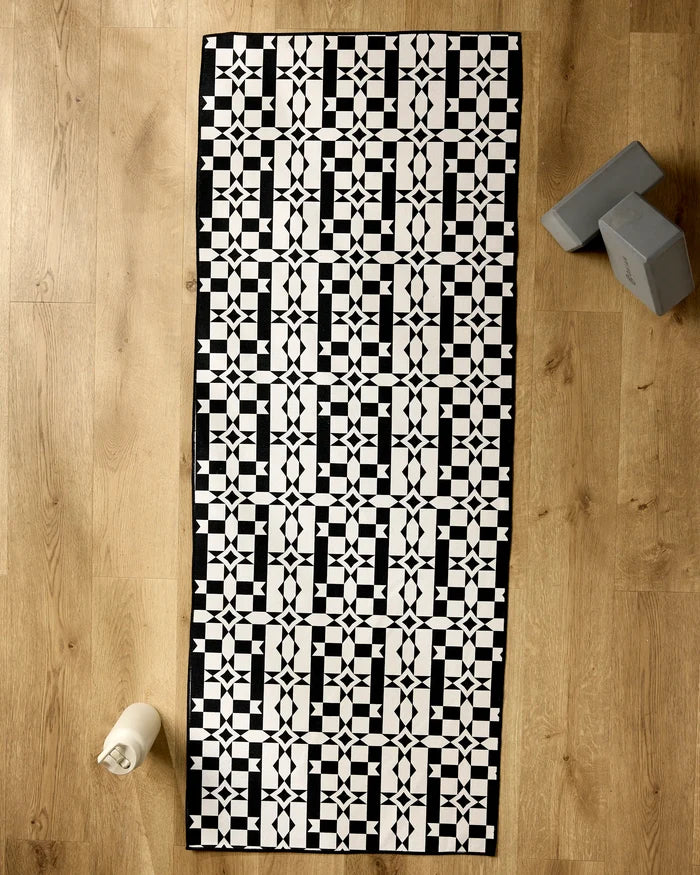 The Geometry Intermission yoga mat with a black and white geometric tiled design is laid out on a wooden floor, accompanied by two gray yoga blocks, a recycled towel, and a white water bottle.