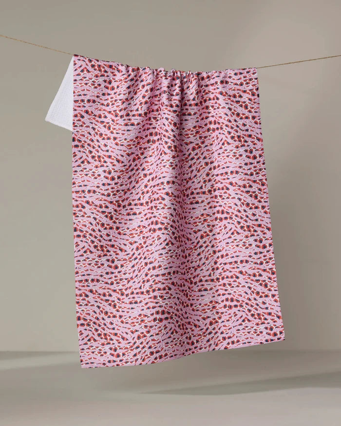 The Happy Hemera "Into the Wild" towel, in pink and white with small red and purple abstract shapes, hangs neatly on a clothesline against a plain, light background.