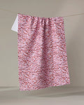 The Happy Hemera "Into the Wild" towel, in pink and white with small red and purple abstract shapes, hangs neatly on a clothesline against a plain, light background.