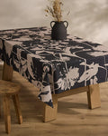 A wooden table draped with the Inverted Floral Black stain-resistant tablecloth from Geometry, topped with a rustic black vase of dried flowers and paired with a simple wooden stool, rests on a light wood floor.