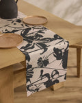 A wooden dining table with a stain-resistant Geometry Inverted Floral table runner, set with simple round wooden plates and a dark pitcher.