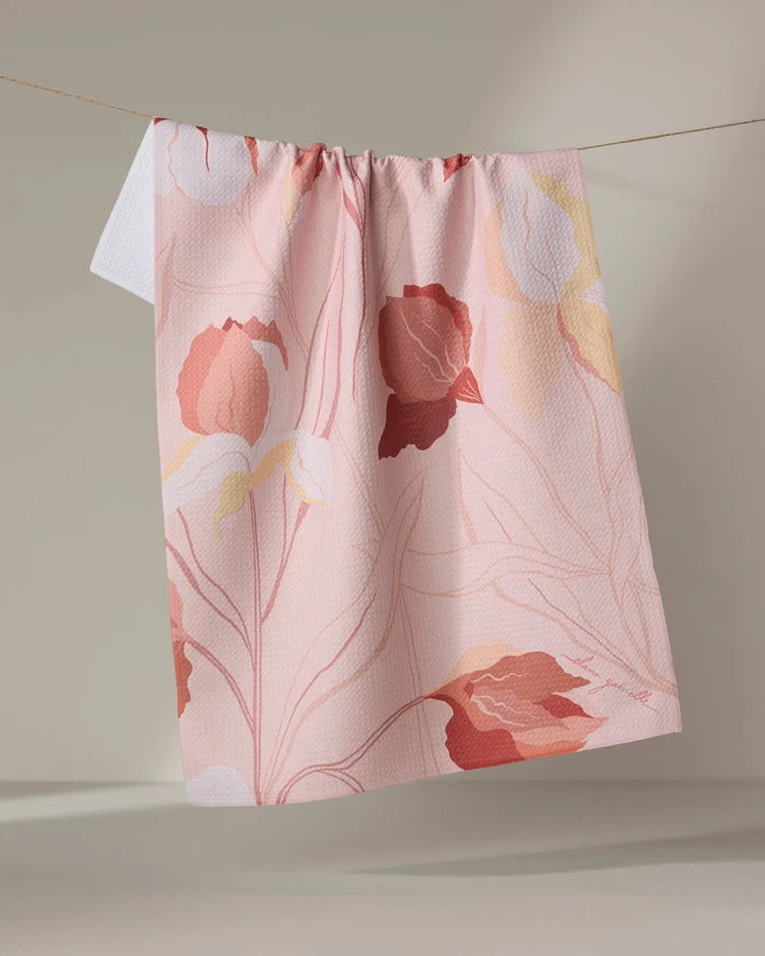 The Iris dish towel by Elana Gabrielle, featuring a large floral pattern in red, pink, yellow, and white on light pink, hangs on a string against a neutral background.