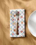 A fork rests on a folded Geometry Italian Summer Grid dinner napkin, featuring a blue grid and cherry pattern, placed on a wooden table next to a white plate.