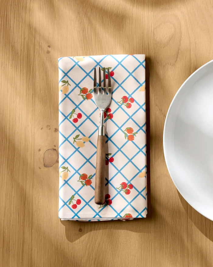 A fork rests on a folded Geometry Italian Summer Grid dinner napkin, featuring a blue grid and cherry pattern, placed on a wooden table next to a white plate.