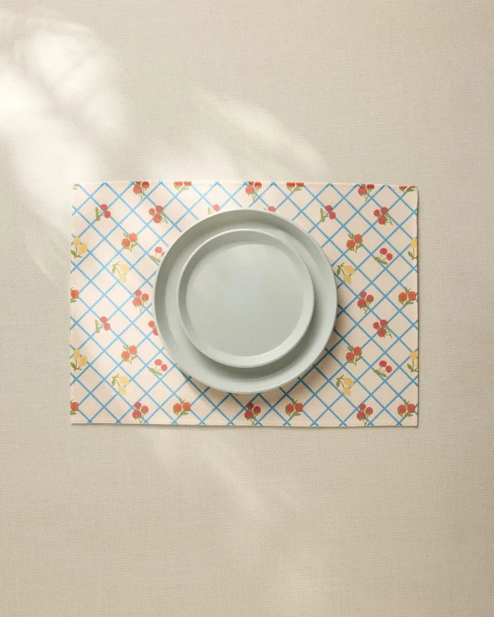 A plain light gray plate sits on the Geometry Italian Summer Grid Placemat, featuring a blue grid and small red and yellow floral pattern, atop a neutral tablecloth.