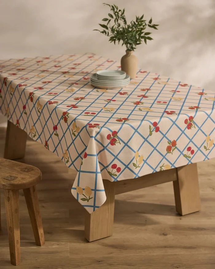 A wooden table is draped with the Geometry Italian Summer Grid tablecloth, showcasing blue grid lines and red and yellow flowers, set with plates and a vase of green foliage. Nearby, a wooden stool rests on a wooden floor.