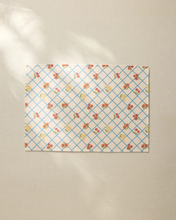 The Italian Summer Grid by Geometry, a rectangular sheet with a blue grid and small floral pattern, rests on a beige textured surface, its recycled placemat style highlighted by soft sunlight and gentle shadows.