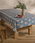 A wooden table is topped with Geometry’s Italian Tiles blue patterned, stain-resistant tablecloth adorned in red and yellow. A beige vase with green leafy branches and a plate of tomatoes sit atop. Nearby is a wooden stool on a light wood floor.