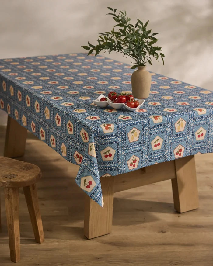 A wooden table is topped with Geometry’s Italian Tiles blue patterned, stain-resistant tablecloth adorned in red and yellow. A beige vase with green leafy branches and a plate of tomatoes sit atop. Nearby is a wooden stool on a light wood floor.